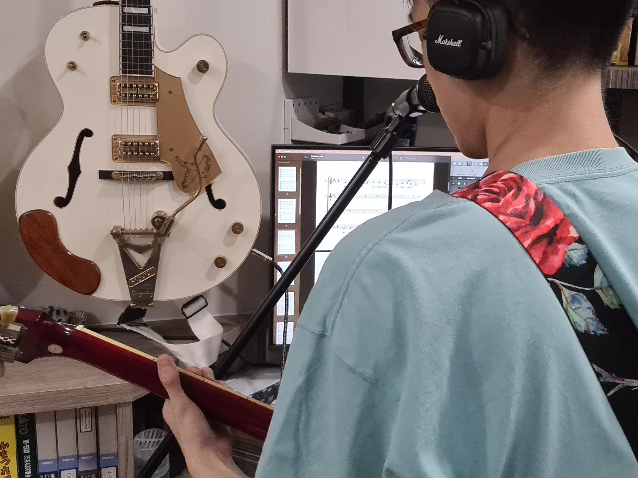 Back shot of John recording vocals while also playing a guitar. There is another hollow-bodied guitar hanging on the wall in front of him.